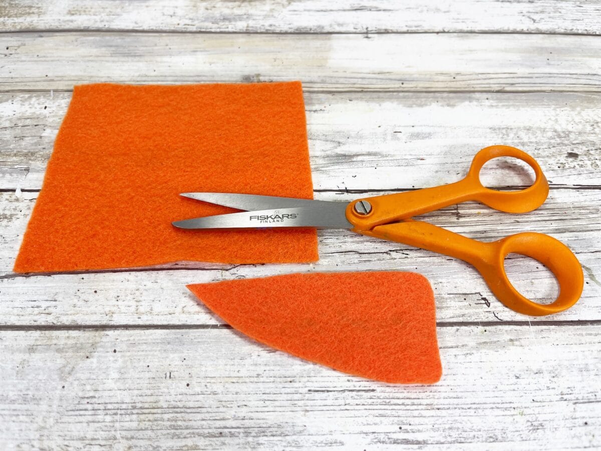 Orange scissors on a wooden surface, next to an orange piece of craft felt, with a cut triangular section.