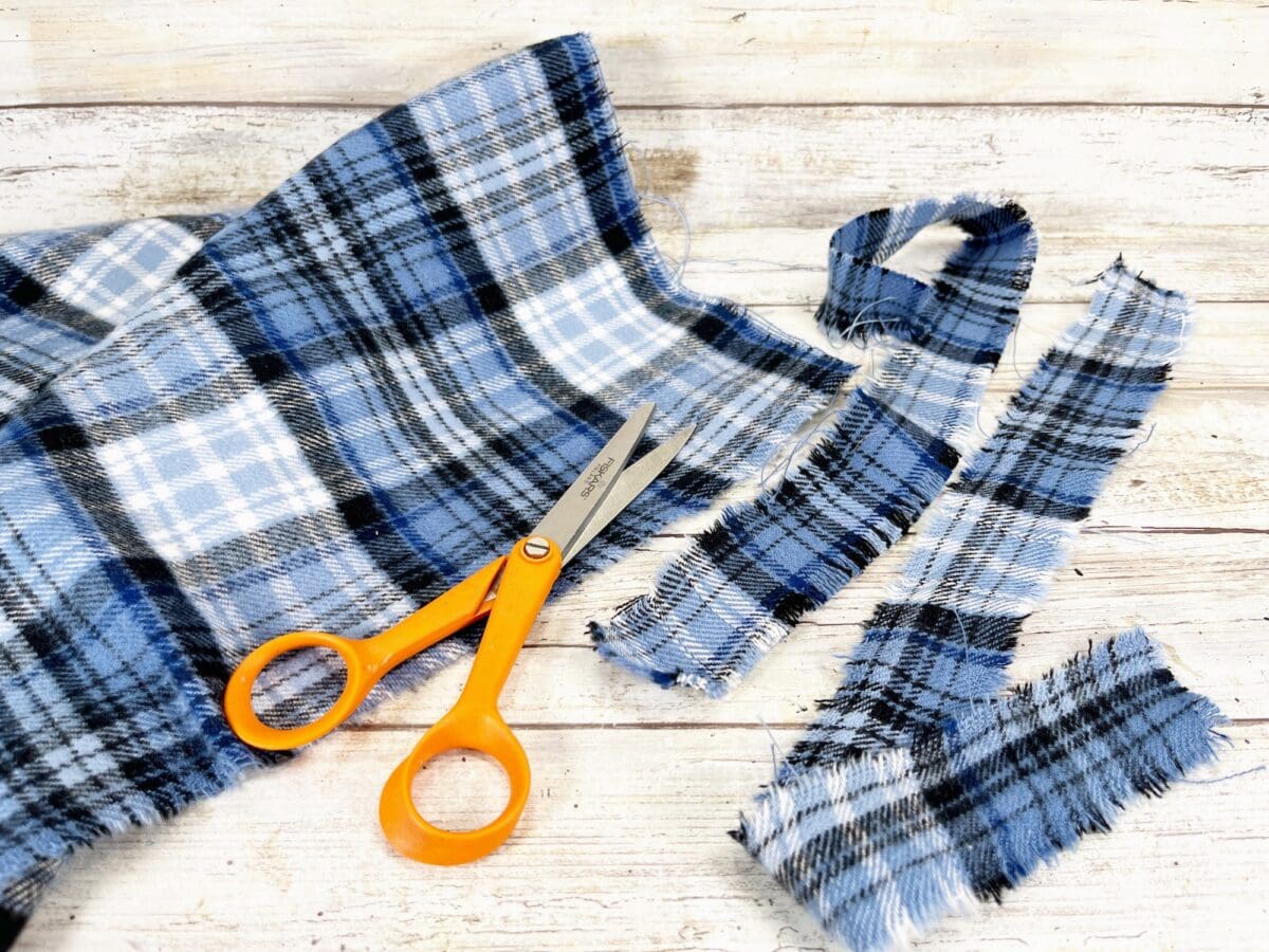 Orange scissors on a wooden surface with cut strips and a larger piece of blue and white plaid fabric.