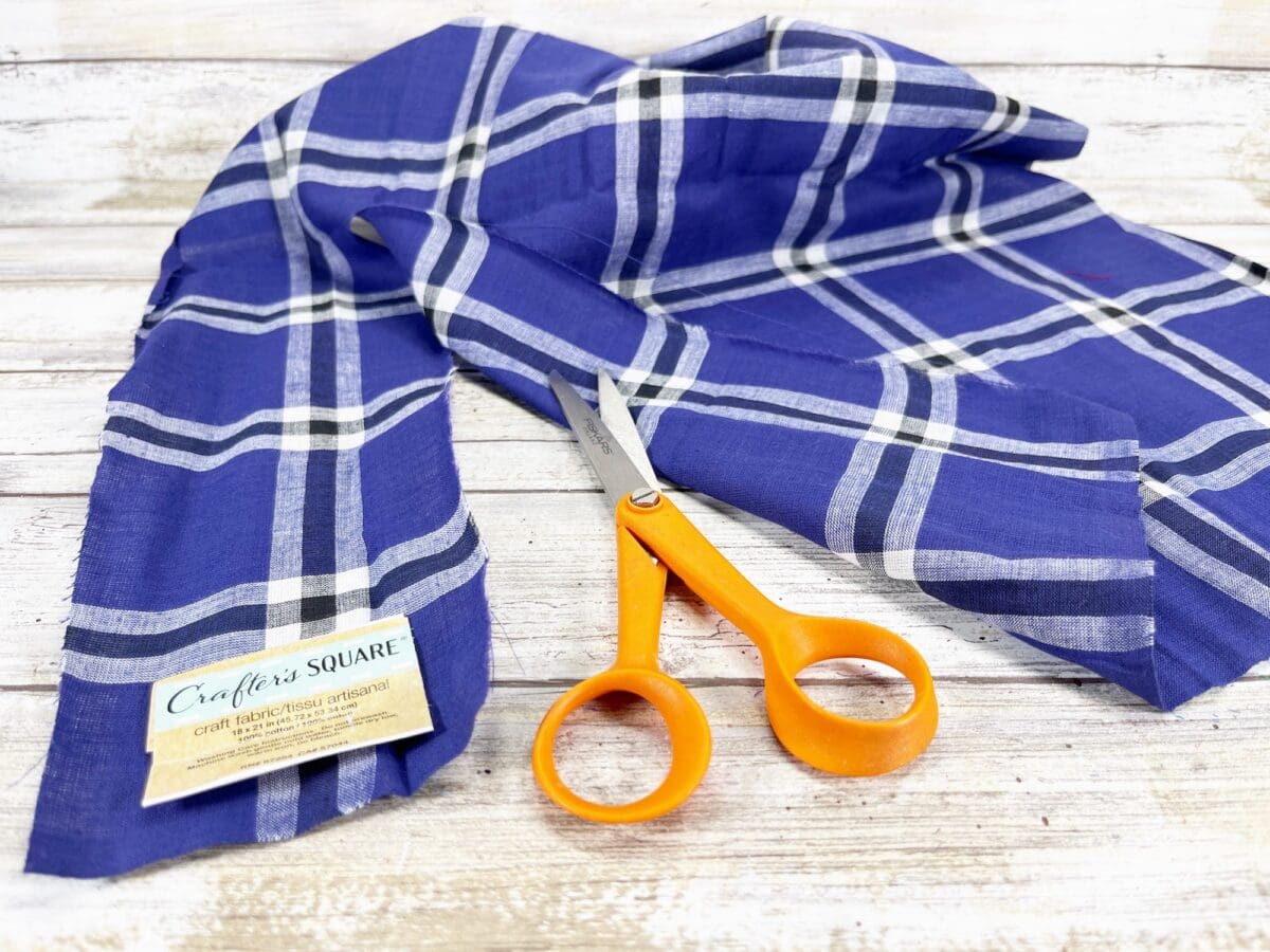 Blue plaid fabric with a label titled "Crafter's Square" is spread out on a wooden surface. An orange pair of scissors rests on the fabric.