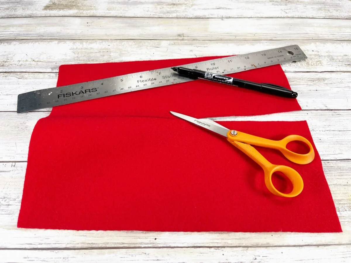 Red fabric piece with an orange-handled scissors, a silver ruler, and a pen placed on a white wooden surface.