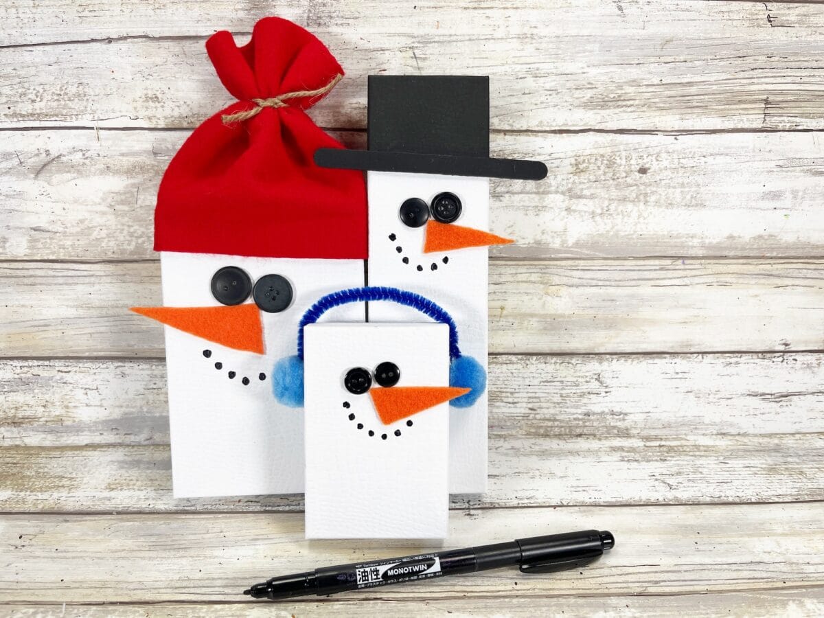 Three rectangular snowmen made from wood blocks with button eyes, orange felt noses, and black marker mouths. One wears a red hat, another a top hat, and the third has blue earmuffs.