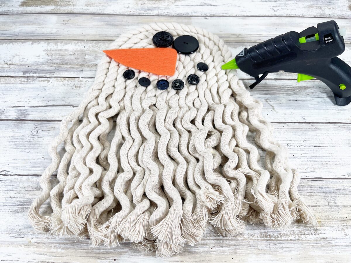 DIY mop ghost being crafted with a glue gun, featuring a mop head, buttons for eyes and mouth, and an orange felt triangle for a nose on a wooden background.