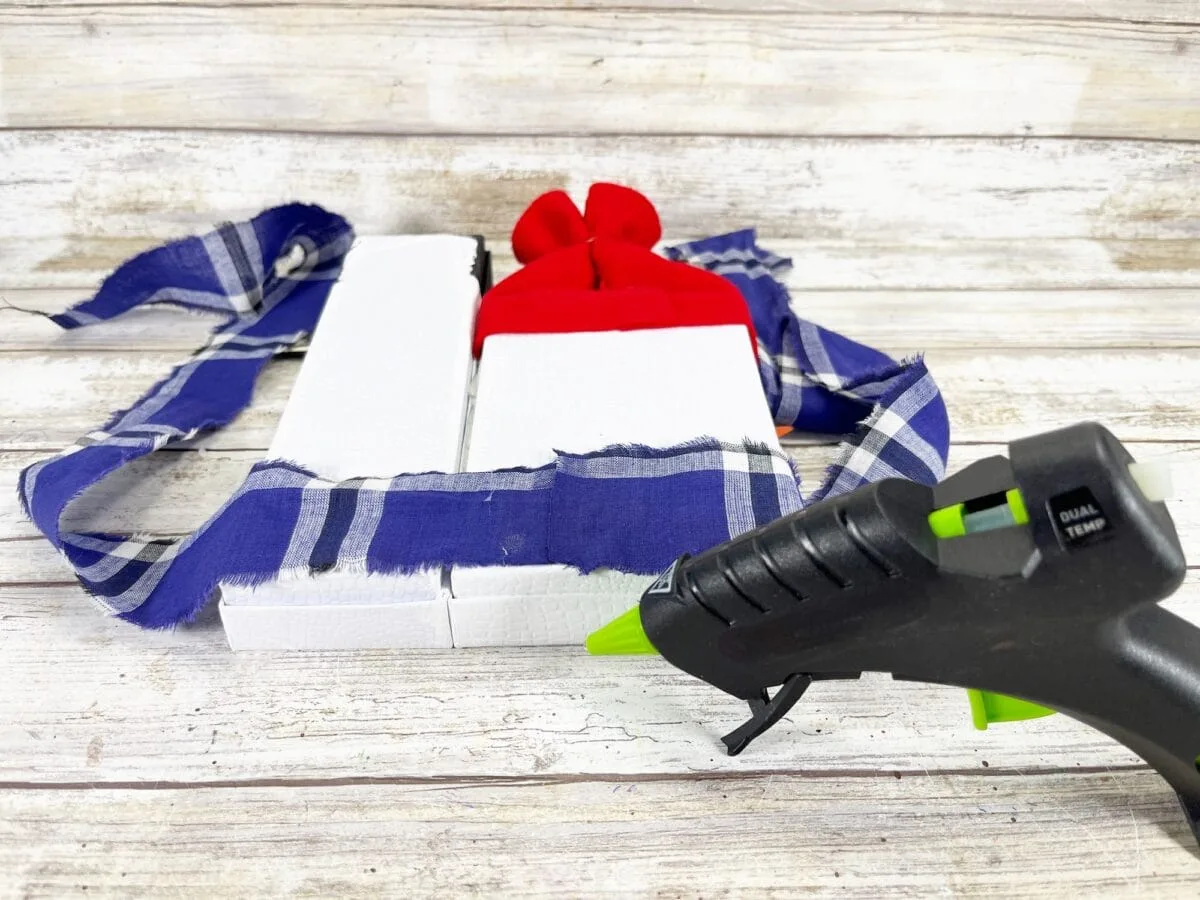 A hot glue gun rests on a wooden surface, with two white rectangular blocks, a red bow, and a torn blue plaid fabric nearby.