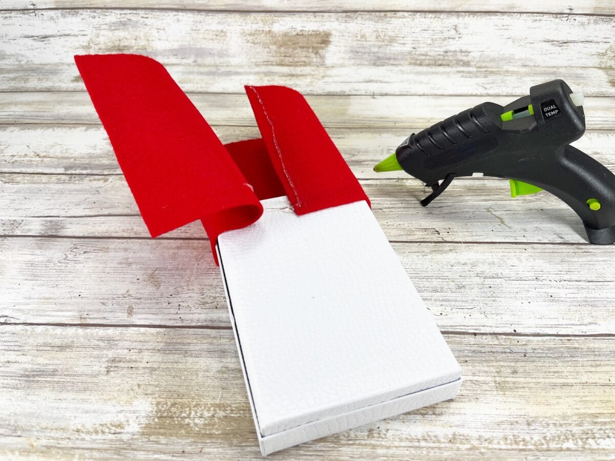 A hot glue gun and red felt sheets are placed on a white textured box, positioned on a wooden surface.