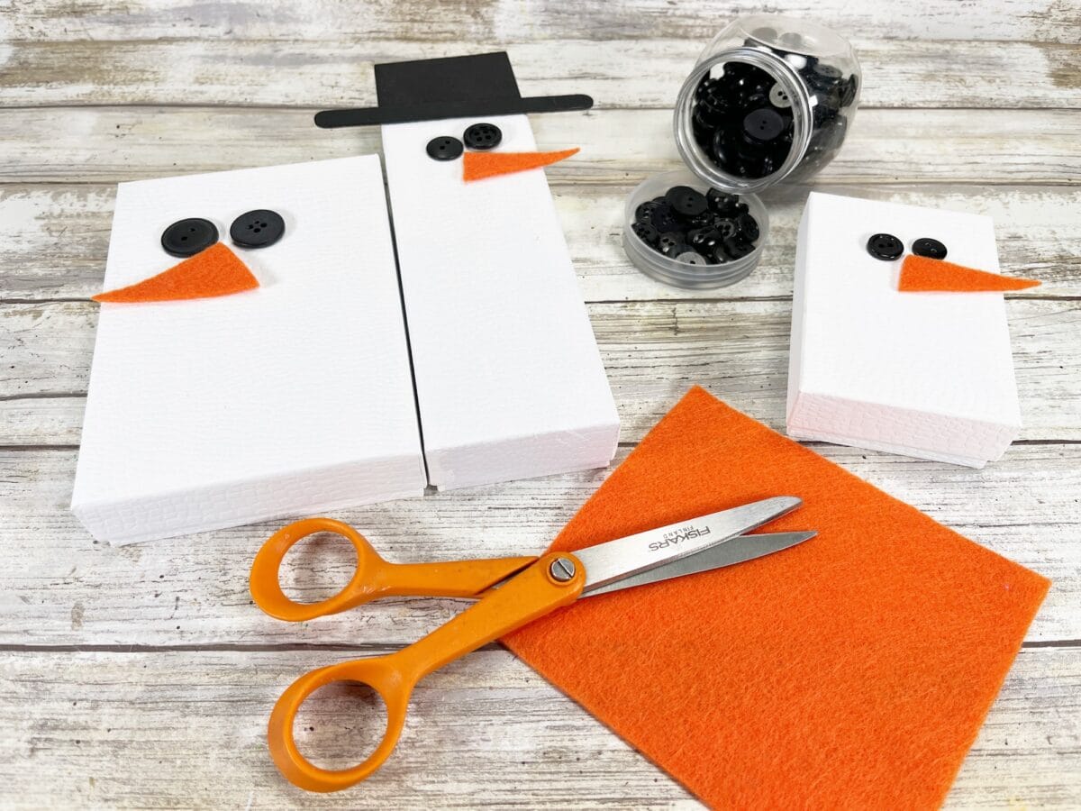 Craft supplies for making snowmen, including white boxes, black buttons, orange felt, scissors, and a black paper hat on a wooden surface.