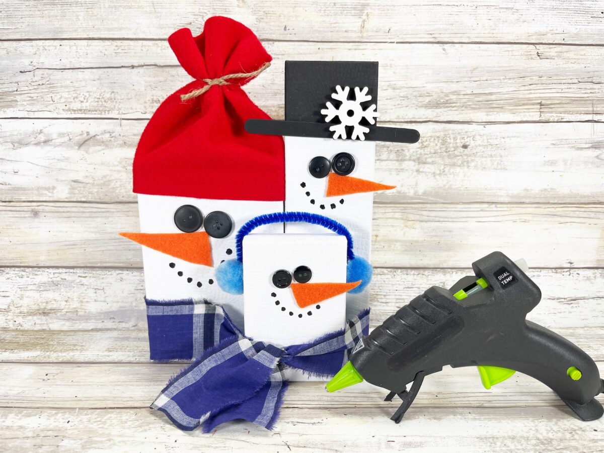 Three snowman-shaped gift boxes adorned with buttons, scarves, and hats are displayed next to a hot glue gun on a wooden surface.