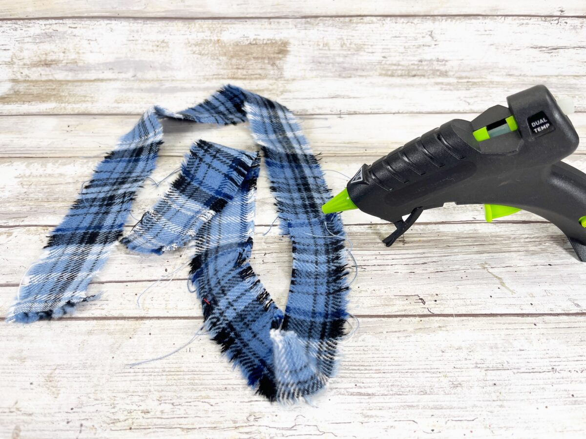 A black glue gun with a green nozzle is beside two strips of frayed blue and black plaid fabric on a rustic wooden surface.