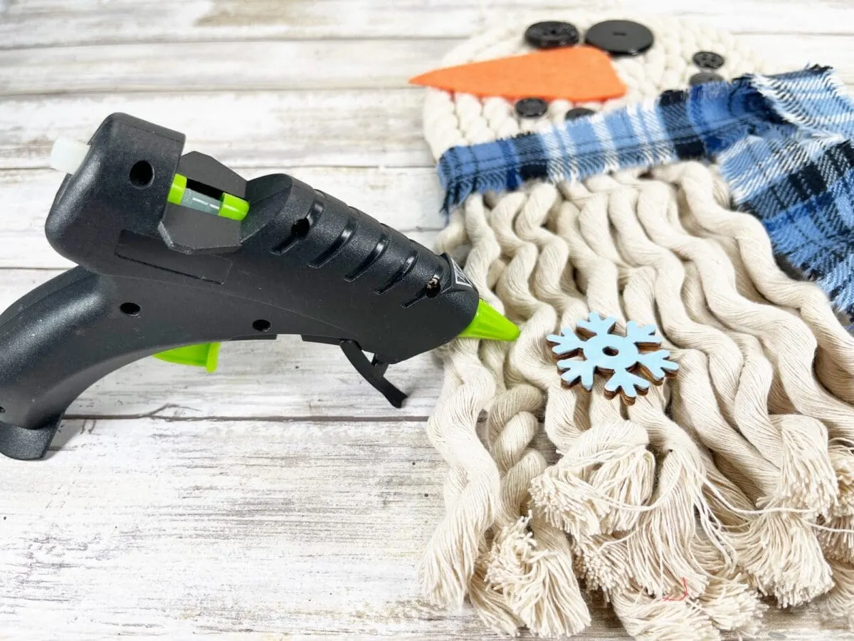 A glue gun is placed on a wooden surface next to a crafting project. The project includes yarn, an orange felt shape, black buttons, and a blue and white plaid fabric piece.