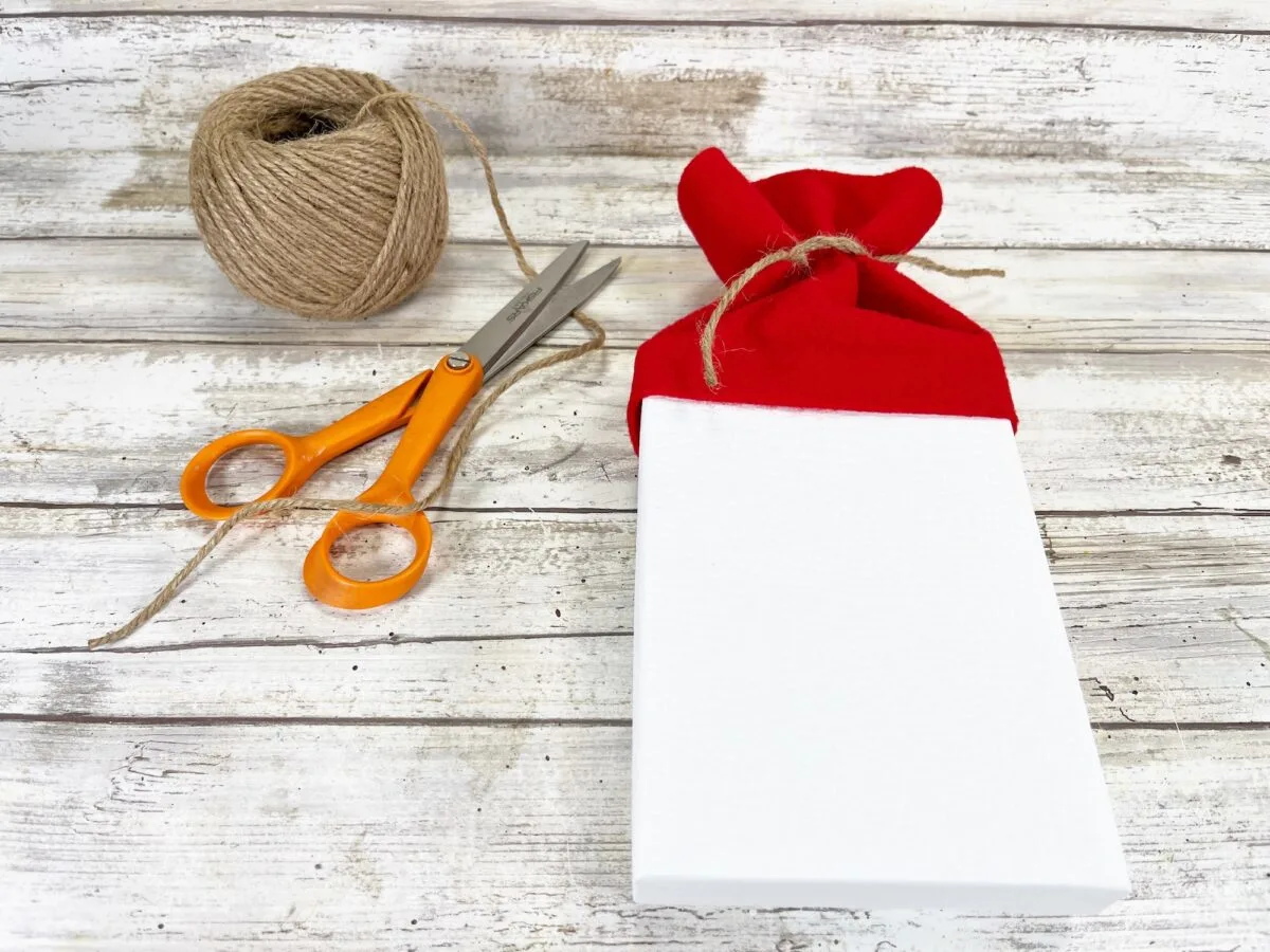 A white gift box with a red cloth covering, tied with twine, next to orange scissors and a ball of twine on a wooden surface.