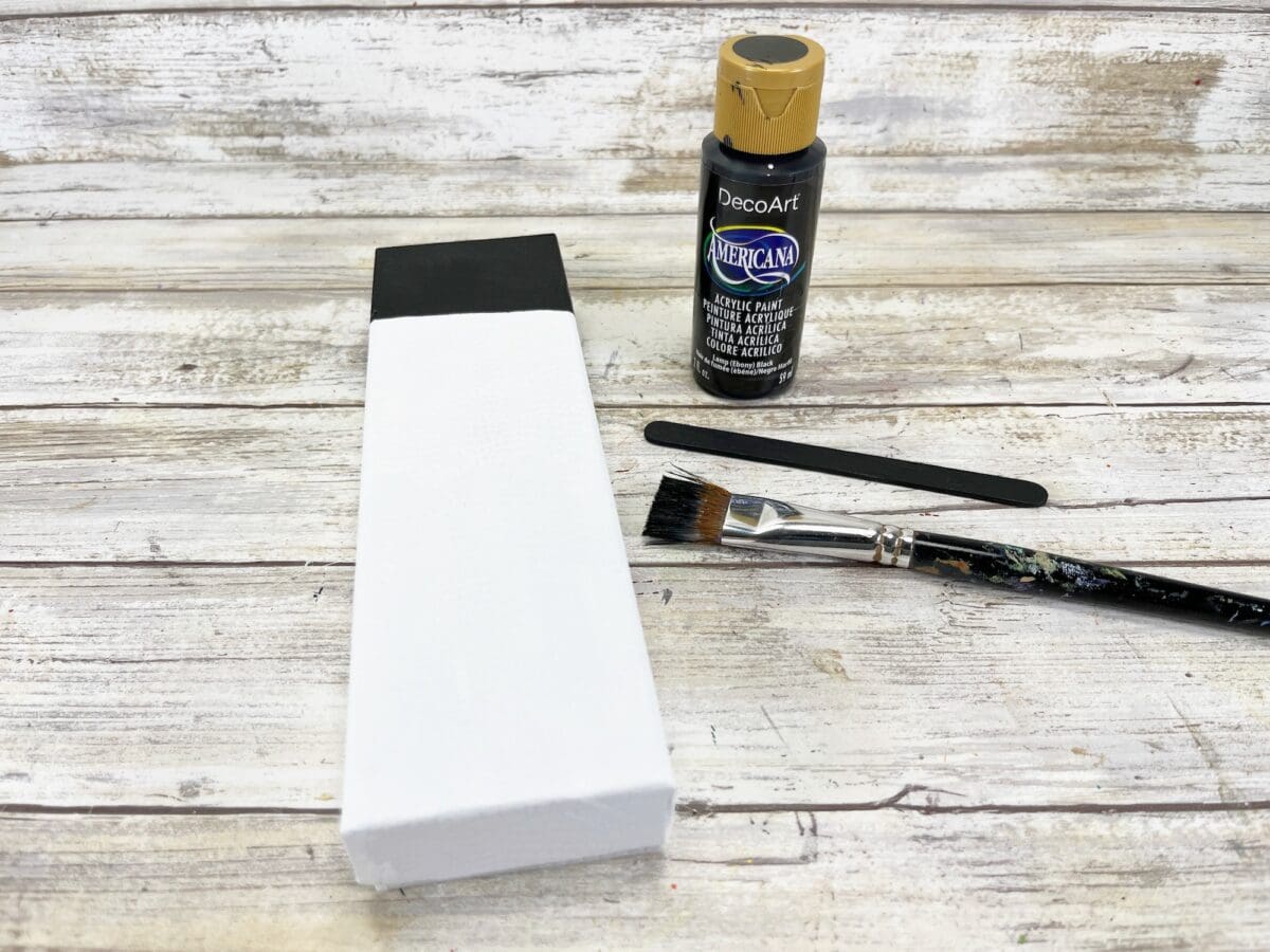 A small white canvas with a black top border, a bottle of black acrylic paint, a flat paintbrush, and a stir stick are on a rustic wooden surface.