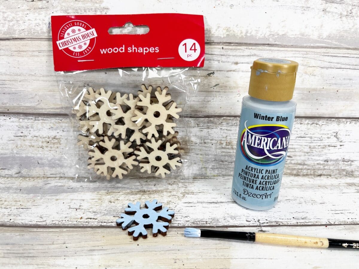 Packet of wooden snowflake shapes, a bottle of blue acrylic paint labeled "Winter Blue," and a paintbrush on a wooden surface. One snowflake is partially painted blue.