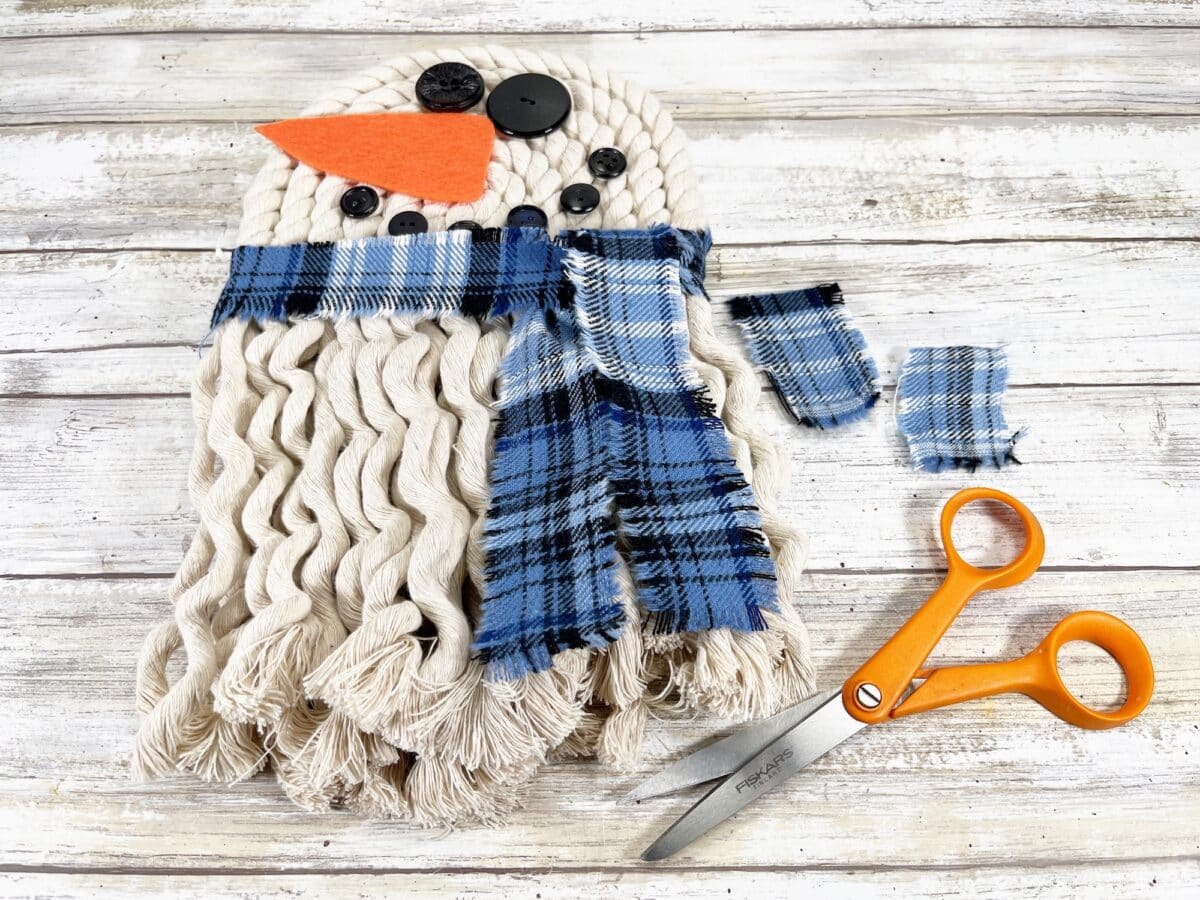 A snowman craft made from braided rope, with a plaid scarf, black button eyes, and an orange nose, lies on a wooden surface next to orange-handled scissors and fabric pieces.