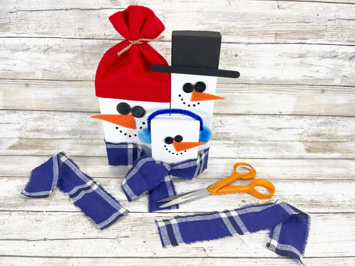 Three block snowmen with fabric accessories and a pair of orange scissors on a white wooden surface.