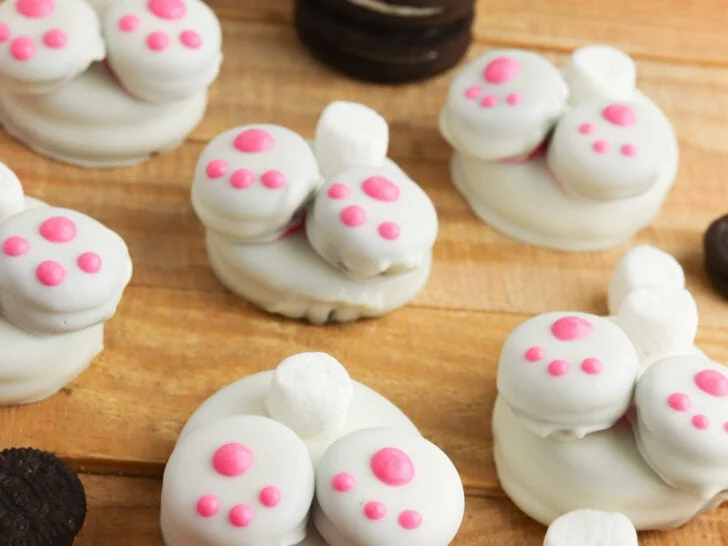 Cookies decorated to resemble bunny feet with pink pads, surrounded by small marshmallows and pastel-colored chocolate eggs on a wooden surface.