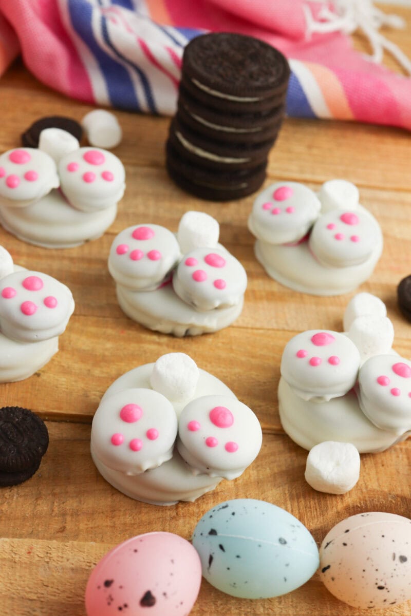 Cookies decorated to resemble bunny feet with pink pads, surrounded by small marshmallows and pastel-colored chocolate eggs on a wooden surface.