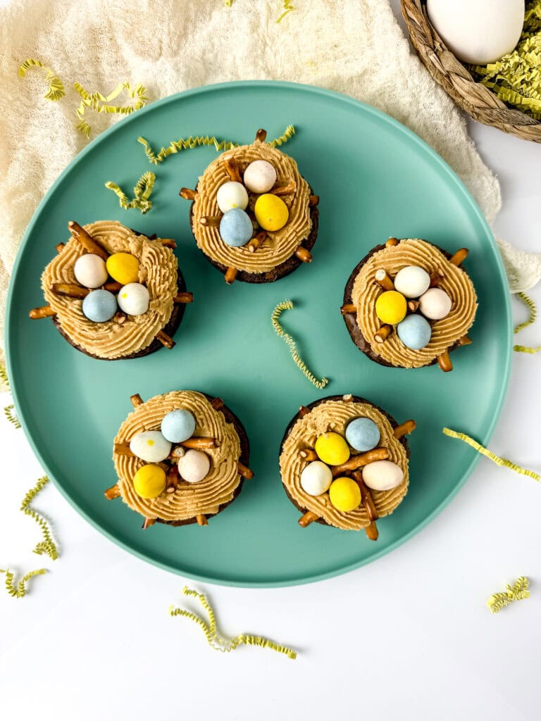 Five cupcakes topped with tan frosting, small chocolate eggs, and pretzel sticks are arranged on a teal plate, resembling delightful bird's nest brownies. Yellow and green paper shreds are scattered around the plate for a playful touch.