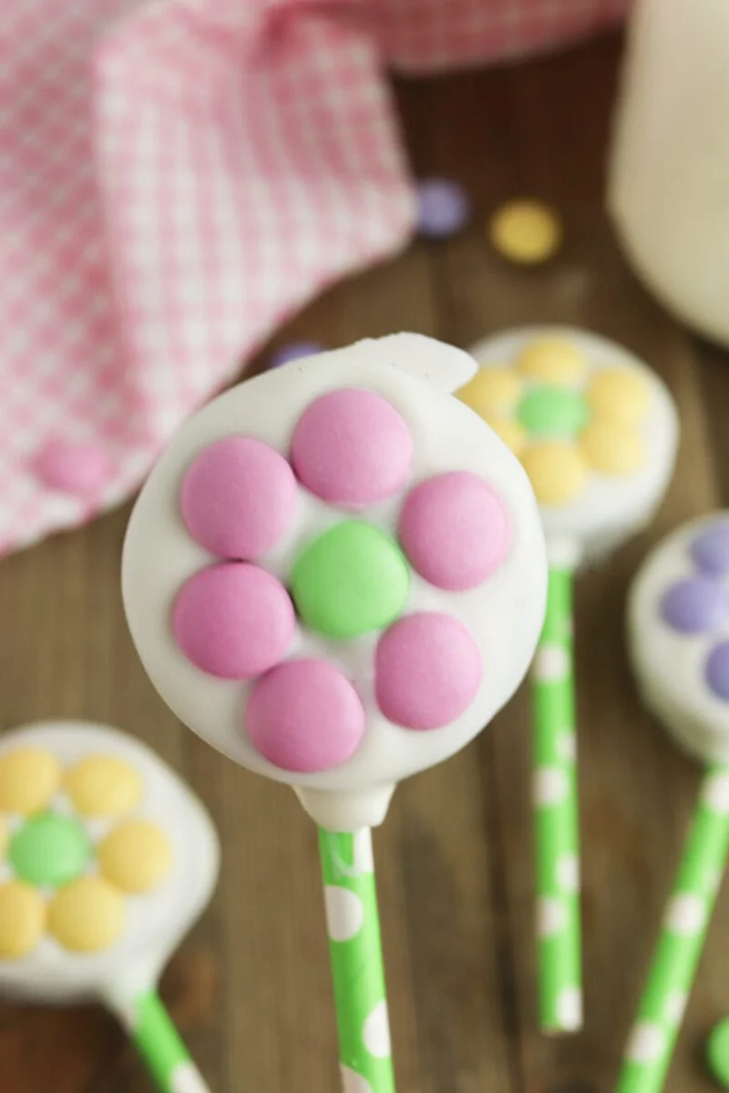 A cookie pop decorated with purple and green candy pieces in a flower shape on a green polka dot stick.