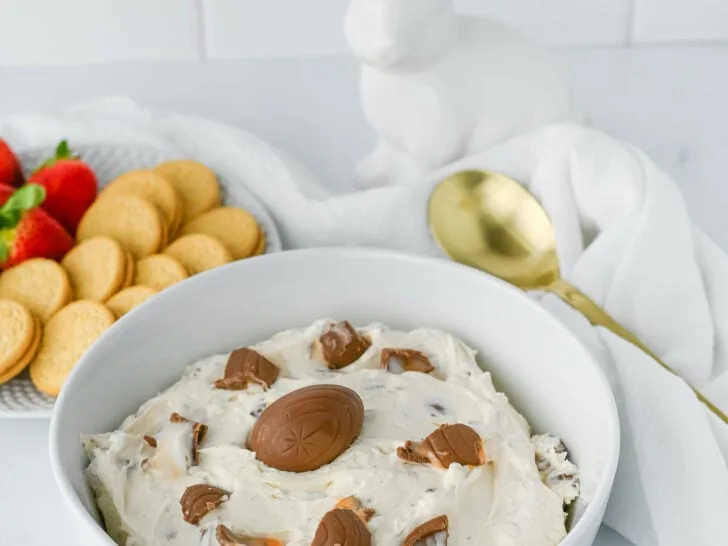 A bowl of dessert dip with chocolate pieces, surrounded by strawberries and cookies. A gold spoon and a white ceramic rabbit are in the background.