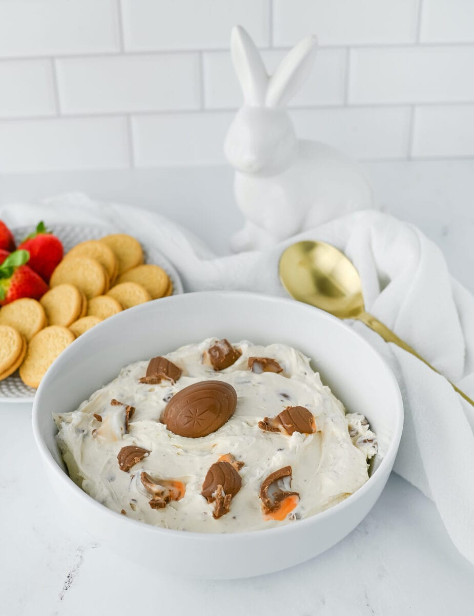 A bowl of dessert dip with chocolate pieces, surrounded by strawberries and cookies. A gold spoon and a white ceramic rabbit are in the background.