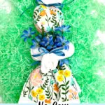 This charming decorative bunny, crafted from a floral dish towel and adorned with blue flowers and ribbons, rests gracefully on green shredded paper. Text reads "SingleGirlsDIY.com" and "No-Sew Dish Towel Bunny," celebrating the delightful art of creating a dish towel bunny.