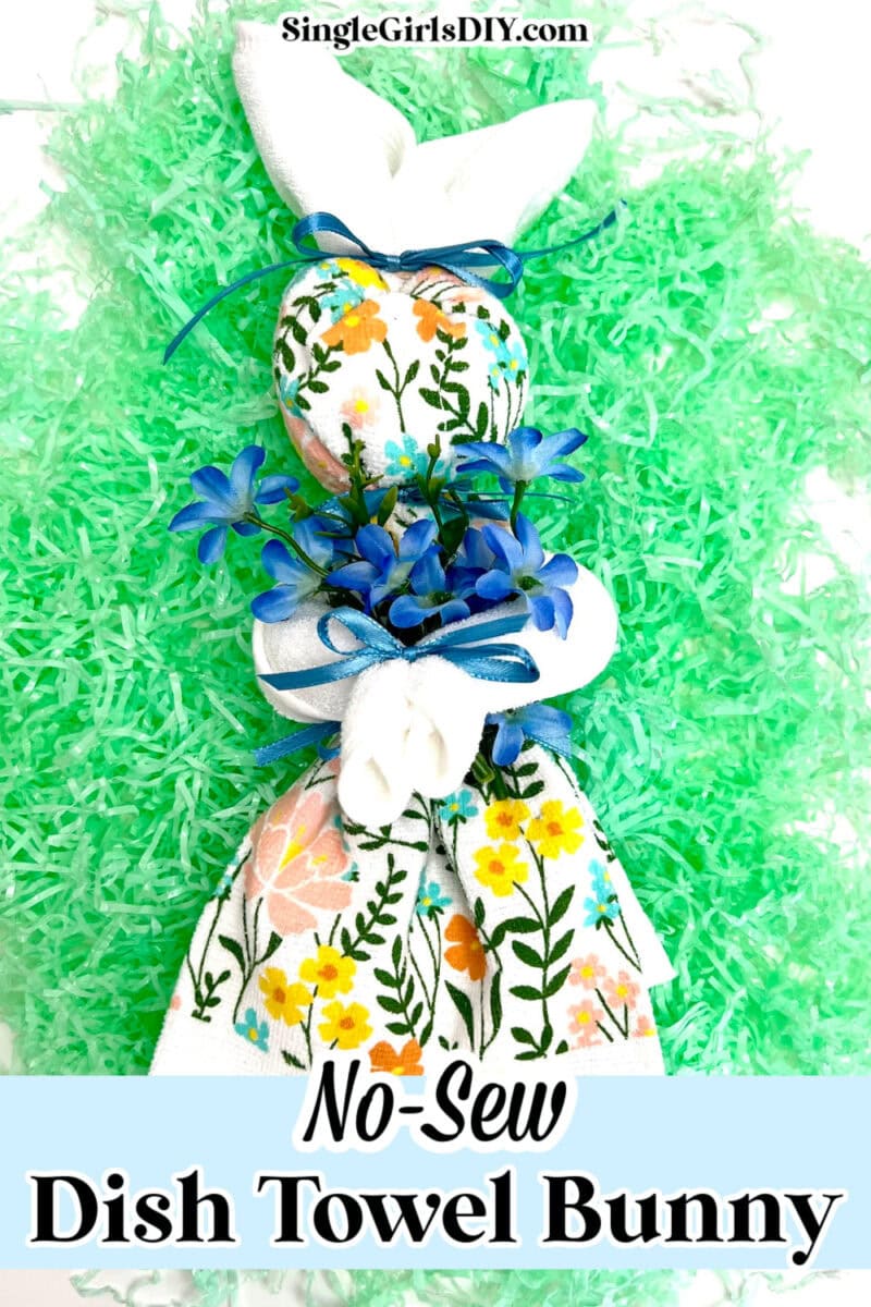 This charming decorative bunny, crafted from a floral dish towel and adorned with blue flowers and ribbons, rests gracefully on green shredded paper. Text reads "SingleGirlsDIY.com" and "No-Sew Dish Towel Bunny," celebrating the delightful art of creating a dish towel bunny.