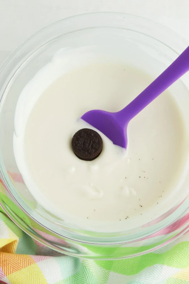 A chocolate cookie is partially submerged in a bowl of white liquid, likely milk, with a purple spatula nearby. A colorful cloth is visible at the bottom.