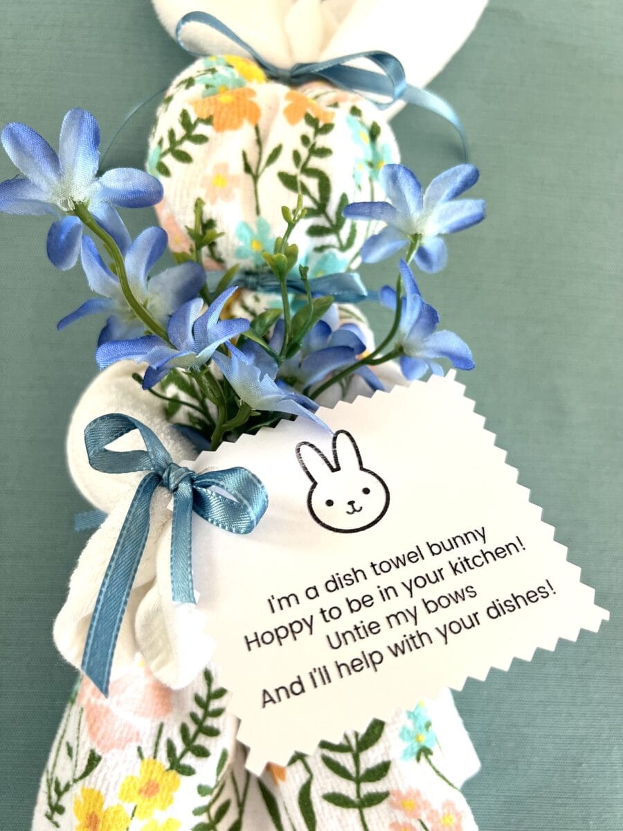 A charming dish towel bunny folded with floral patterns, adorned with blue flowers and a ribbon, carries a note featuring a whimsical bunny illustration and playful message.