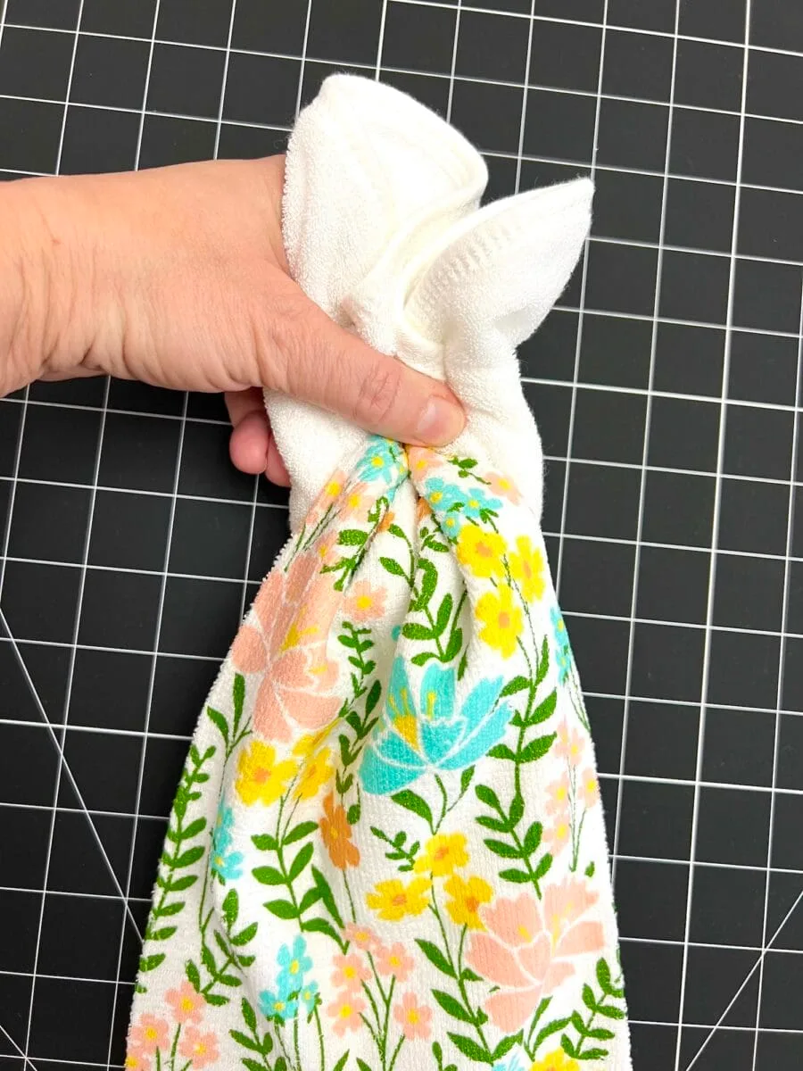 A hand holds a floral-patterned cloth with a grip, against a black grid background. The cloth features pink, blue, and yellow flowers with green leaves.