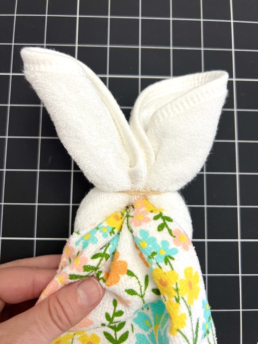 A hand holding a towel folded into a bunny shape, with ears at the top and floral patterns at the bottom, set against a black grid background.