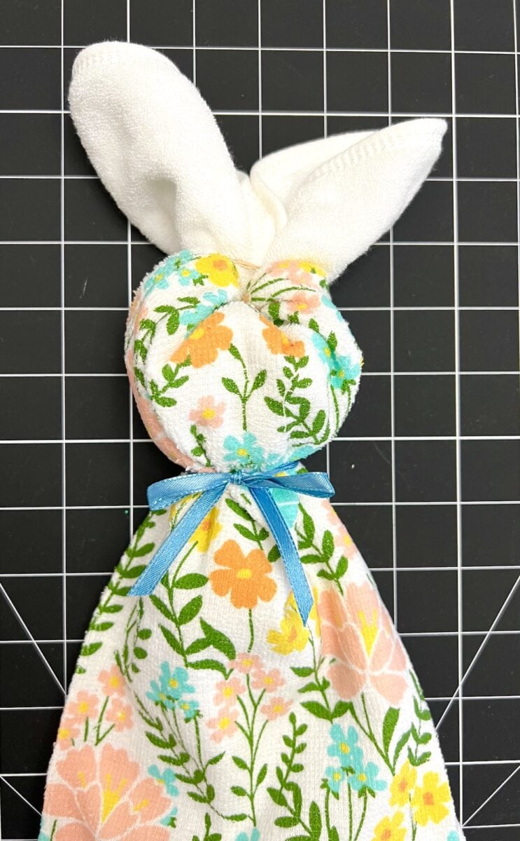 Fabric crafted into a bunny shape with floral and leaf patterns tied with a blue ribbon, on a grid-lined black background.