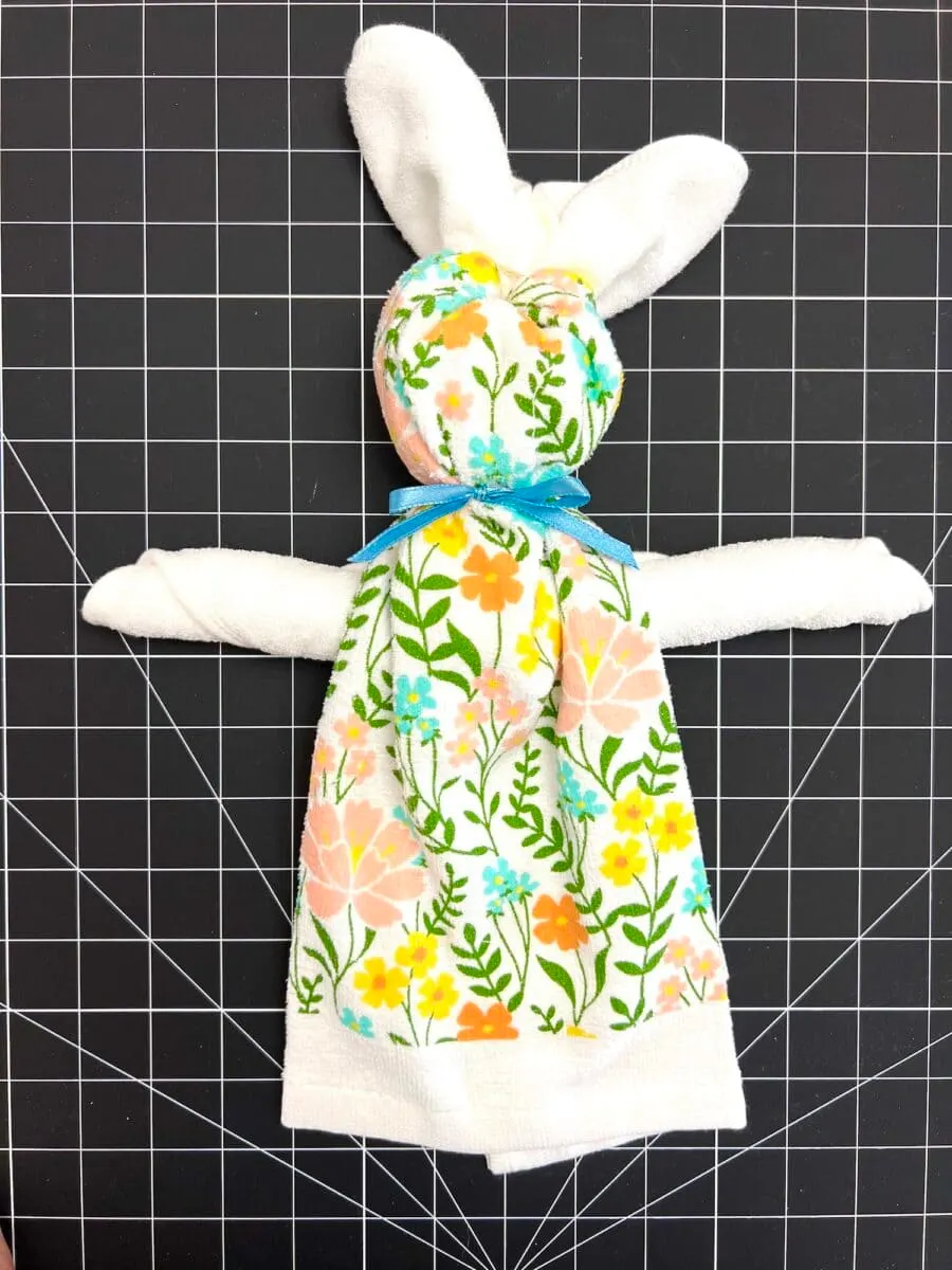 A fabric bunny, reminiscent of a dish towel creation, features a floral-patterned body and white limbs resting on a black grid background. A blue ribbon elegantly adorns its neck.