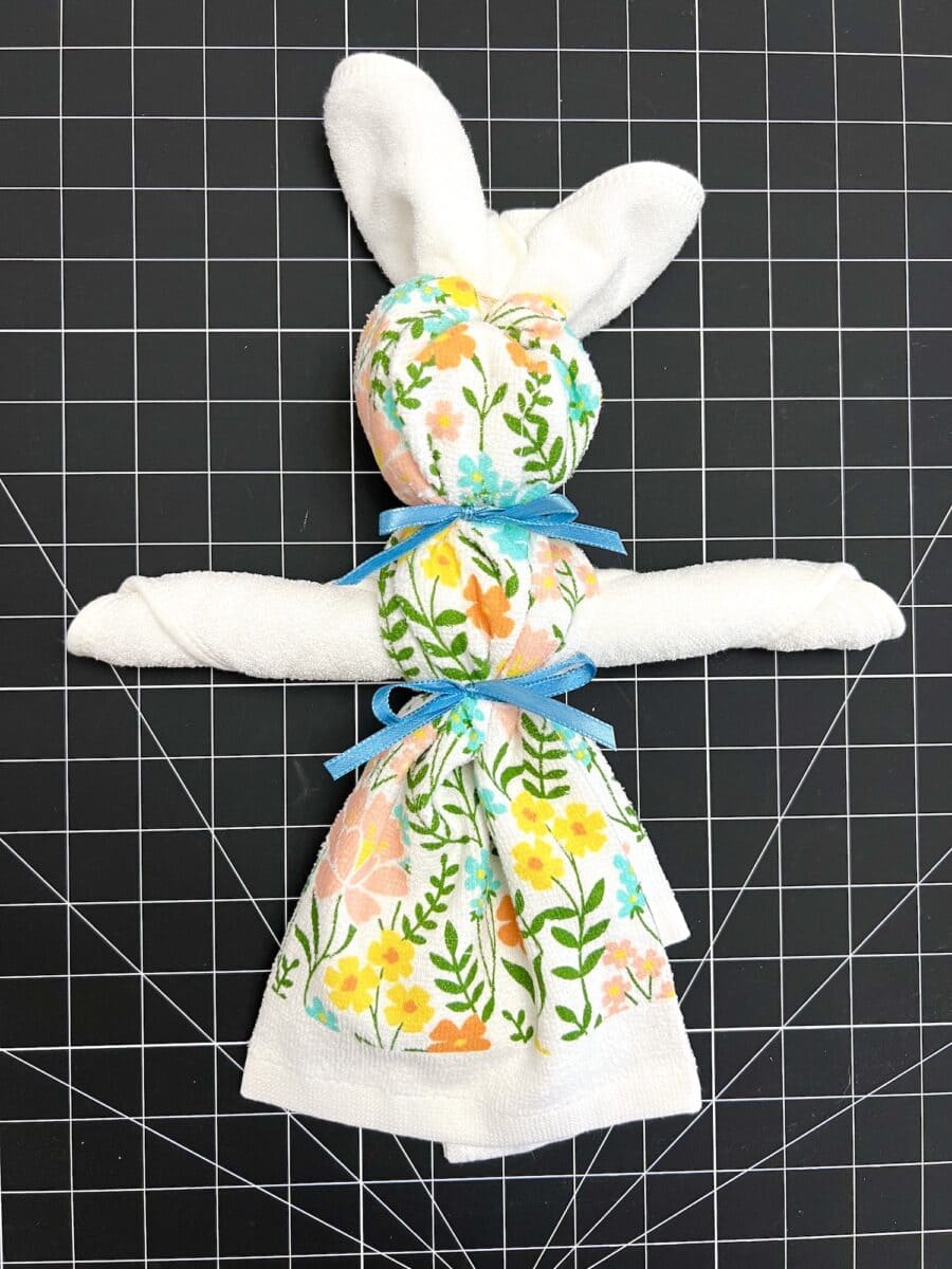 A charming cloth doll, this dish towel bunny is crafted from a floral-patterned towel. It features a blue ribbon around its neck and waist, elegantly displayed on a black grid surface.