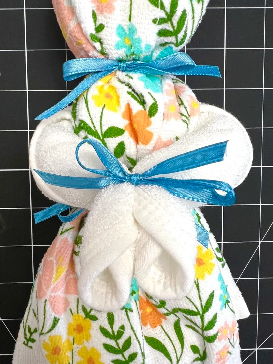 A dish towel bunny folded into a charming bunny shape, adorned with colorful floral patterns and tied with a blue ribbon, sits elegantly against a black grid background.