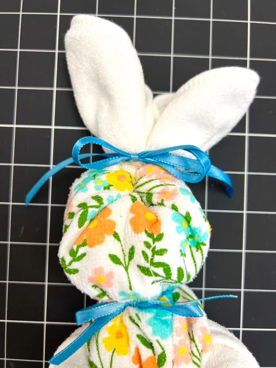 A charming fabric bunny with floral patterns, secured with blue ribbons, is positioned on a grid-patterned dish towel surface.