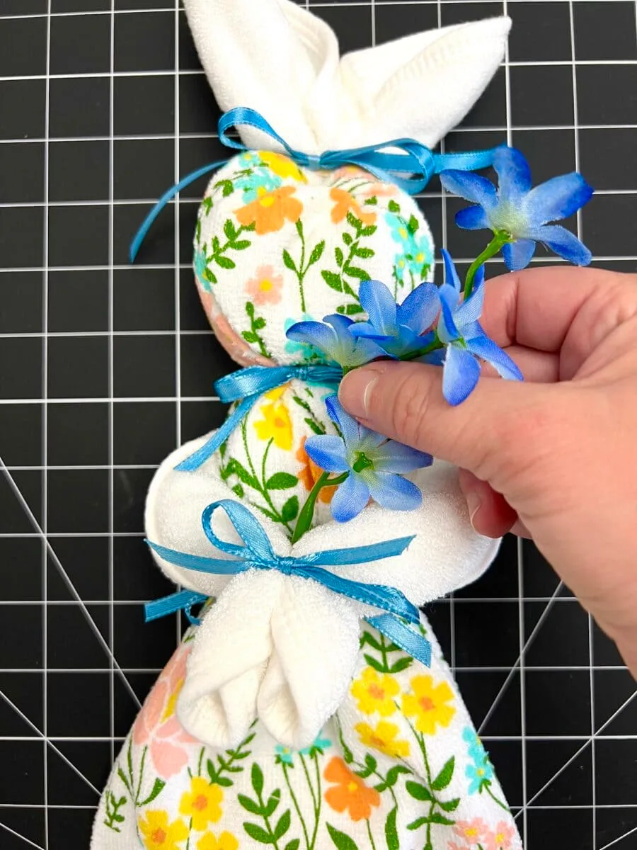 A hand places blue flowers on a decorative cloth bunny, adorned with colorful floral patterns and tied with blue ribbons. This charming scene unfolds against a black grid surface, as if a whimsical dish towel bunny has come to life amidst blooming artistry.