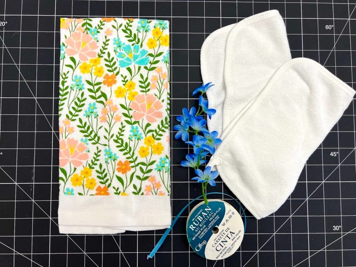 Floral fabric, white cloth pads, blue decorative flowers, and a roll of ribbon on a black grid cutting mat.