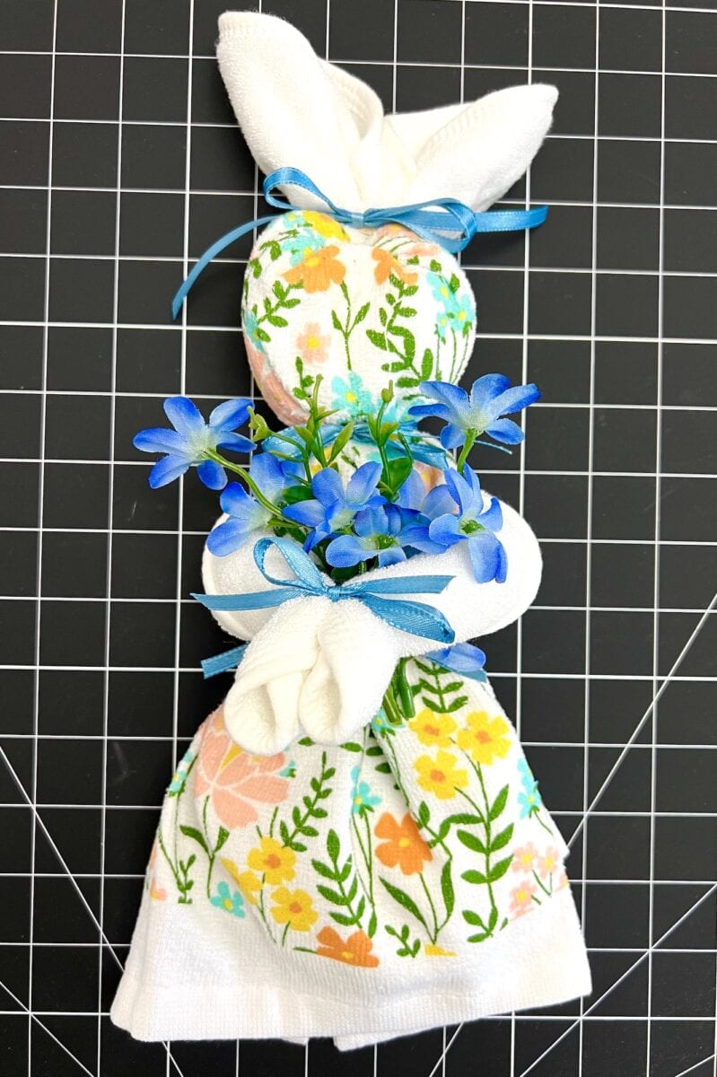 A charming dish towel bunny adorned with flower patterns, blue ribbons, and artificial blue flowers sits gracefully on a black grid surface.