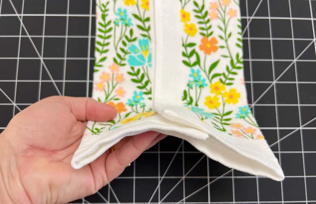 Hand holding a folded, floral-patterned towel with green leaves and colorful flowers on a black grid background.