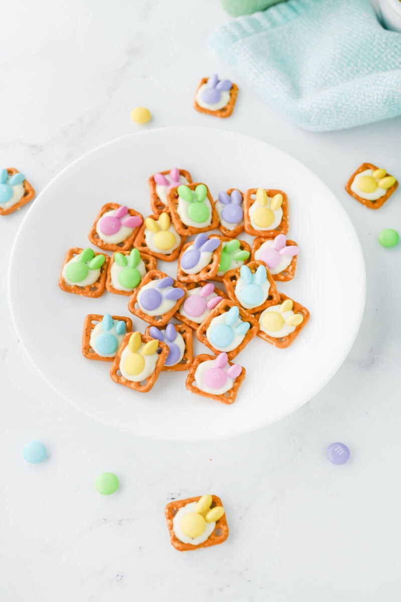 Easter bunny pretzel treats feature square pretzels topped with pastel candy melts in charming bunny shapes, artfully displayed on a white plate, and surrounded by scattered candy and a draped blue cloth.