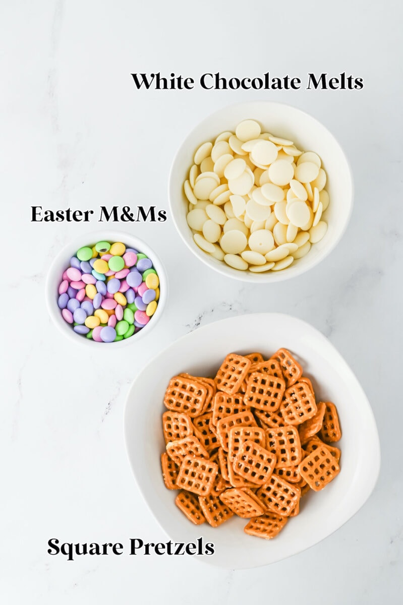 Bowls of white chocolate melts, Easter M&Ms, and square pretzels on a white surface await transformation into delightful Easter bunny pretzel treats.