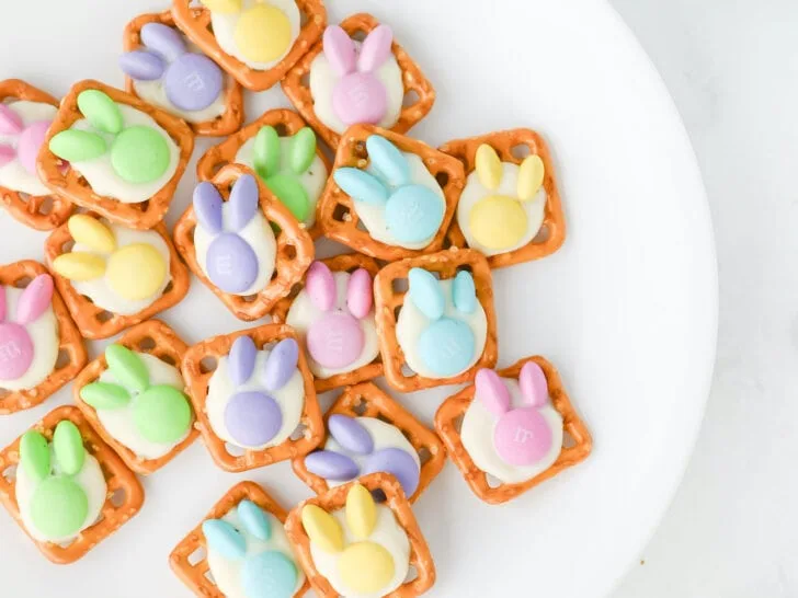 A white plate displays delightful Easter bunny pretzel treats, with squares topped by colorful bunny-shaped candies. Scattered around are extra candies and pretzels, adding a playful touch to the scene.