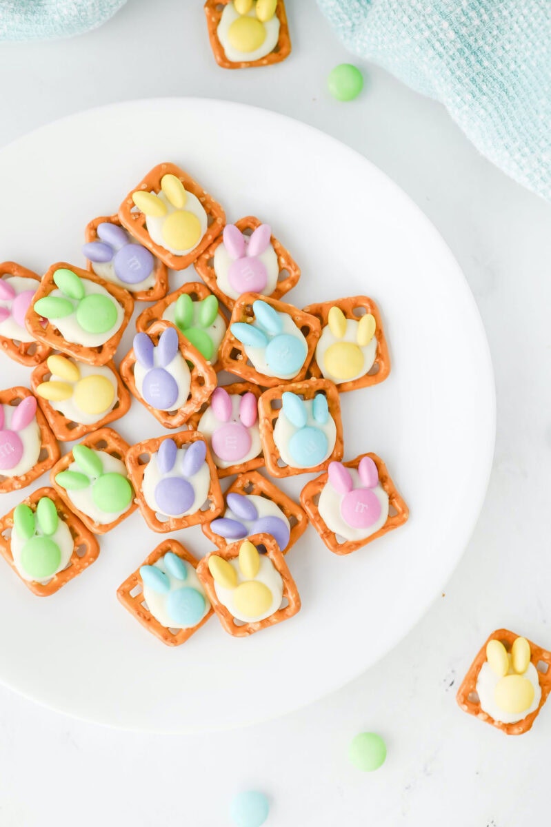 A white plate displays delightful Easter bunny pretzel treats, with squares topped by colorful bunny-shaped candies. Scattered around are extra candies and pretzels, adding a playful touch to the scene.