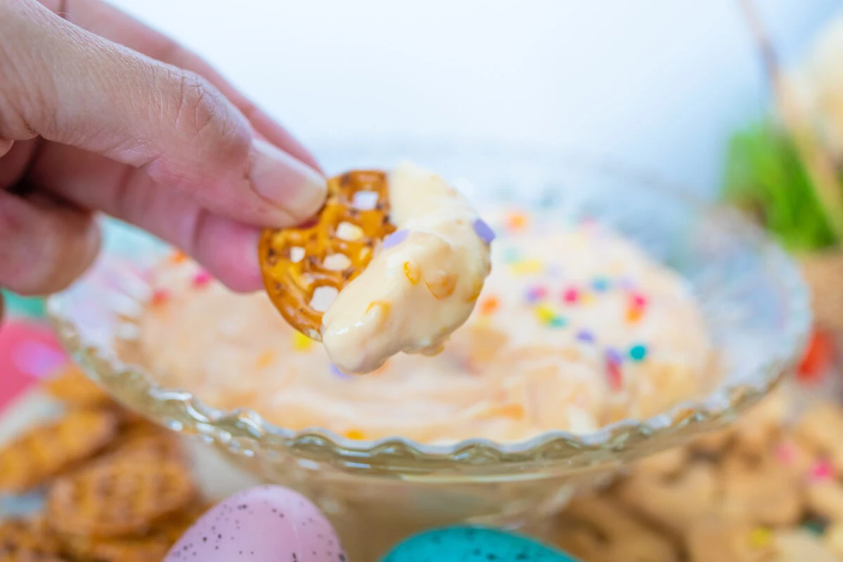 A hand dips a pretzel into a creamy, sweet Easter dip topped with colorful sprinkles, while pastel-colored eggs peek from the background.