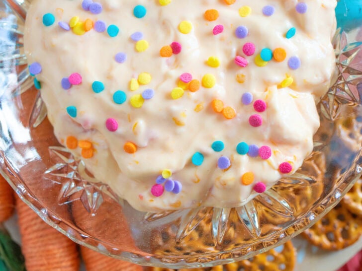 A sweet Easter dip sits in a bowl, topped with colorful confetti sprinkles and surrounded by crunchy pretzels and decorative faux carrots.