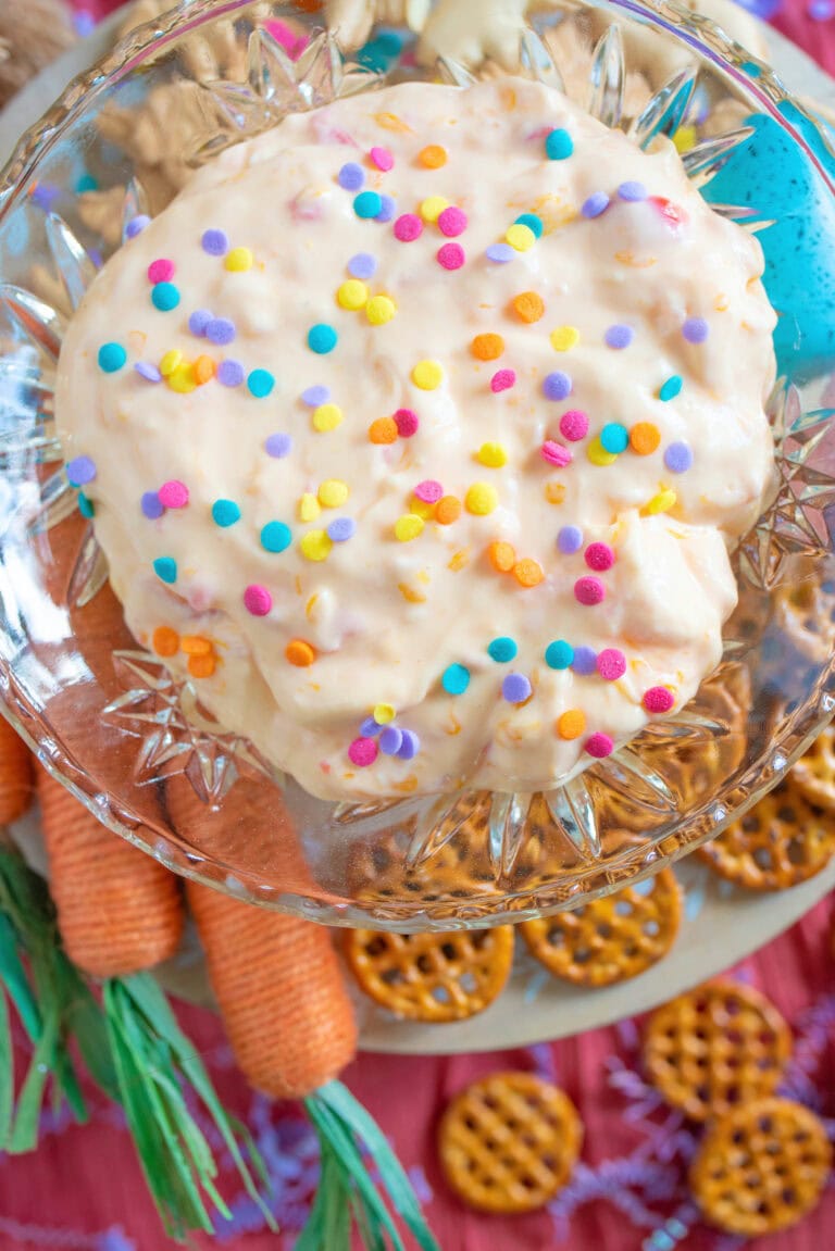 A sweet Easter dip sits in a bowl, topped with colorful confetti sprinkles and surrounded by crunchy pretzels and decorative faux carrots.