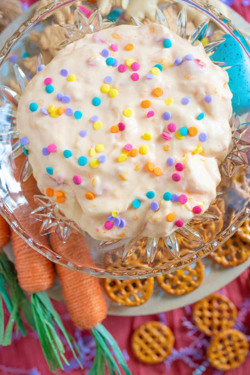 A sweet Easter dip sits in a bowl, topped with colorful confetti sprinkles and surrounded by crunchy pretzels and decorative faux carrots.