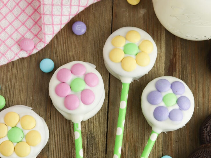 Colorful flower-shaped cookie pops with pastel candies on green sticks, placed on a wooden surface beside a jar of milk, a pink checkered cloth, and scattered chocolate cookies.