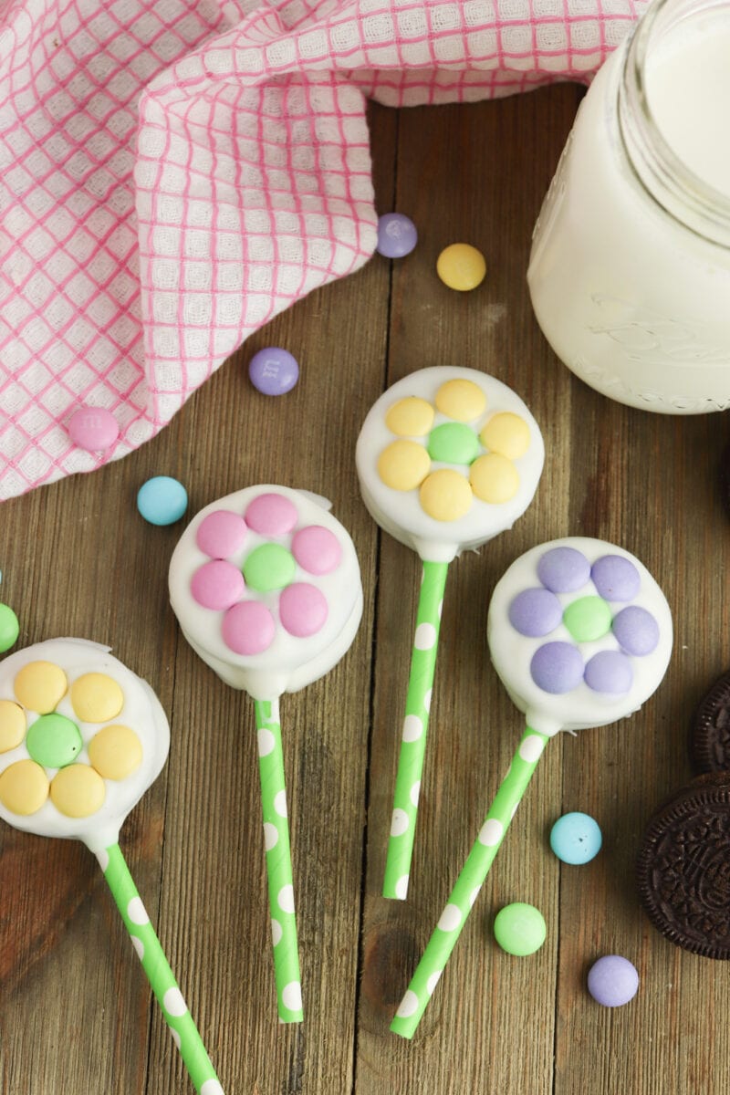 Colorful flower-shaped cookie pops with pastel candies on green sticks, placed on a wooden surface beside a jar of milk, a pink checkered cloth, and scattered chocolate cookies.