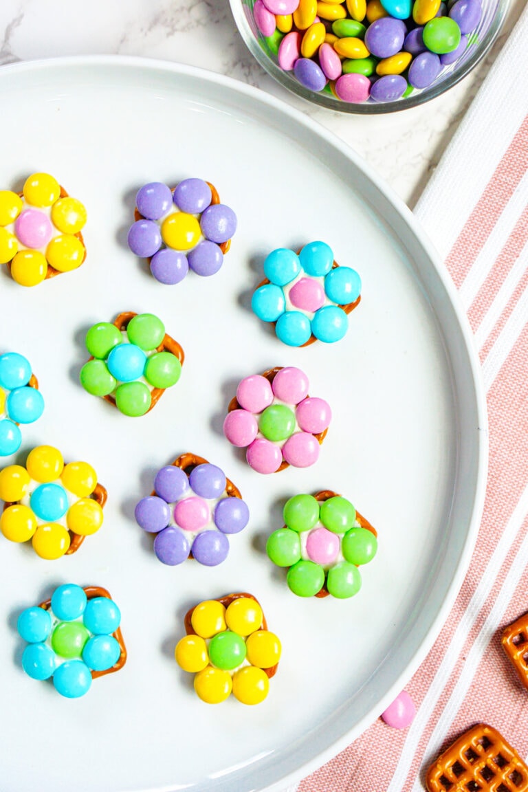 A plate with flower-shaped candy arrangements using pretzels, coated in colorful chocolate pieces, next to a bowl of similar candies and a striped cloth.