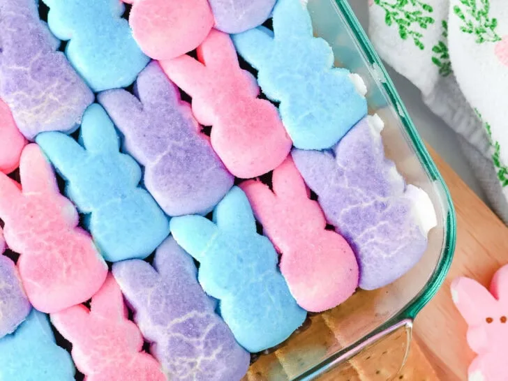 A glass dish containing rows of pink, blue, and purple marshmallow bunnies on top of graham crackers, with a carrot-patterned cloth nearby.